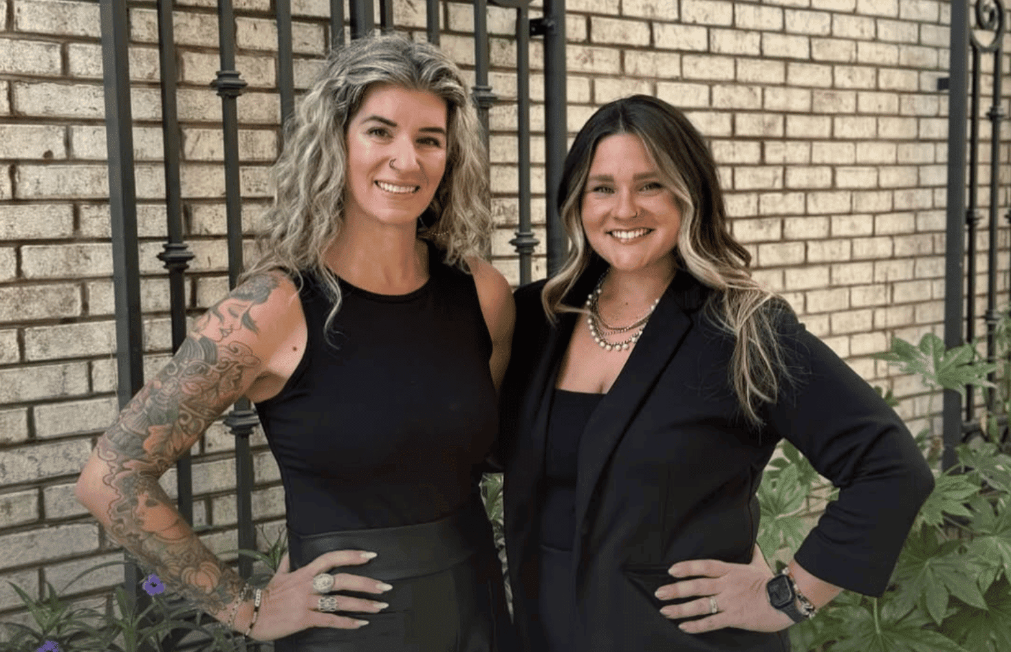 Two women smiling and standing together in front of a brick wall.